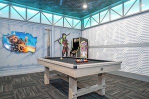 Una mesa de billar en una habitación con techo temático de galaxia, un mural de un personaje y una pared con diseño.