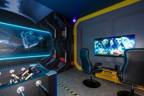 Une salle de jeux équipée d’un baby-foot, de deux fauteuils de jeu et d’un grand téléviseur diffusant un jeu vidéo.