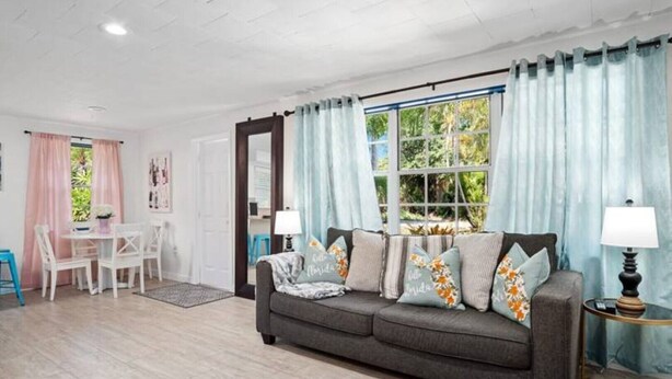 A cozy living room with a grey sofa, a small table, and chairs. There is a window with curtains, a lamp, and a mirror on the wall. - King Bed Near Pickleball, Beach & Downtown (Naples)