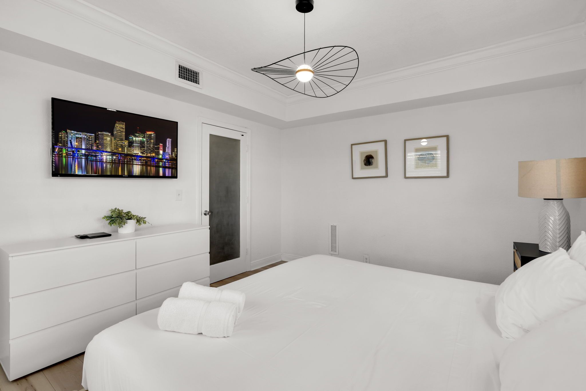 Una habitación de hotel moderna con una cama grande, un televisor de pantalla plana montado en la pared, una lámpara de techo original y una puerta que da a otra habitación.