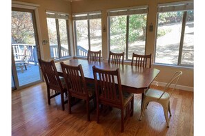 A dining area with a large wooden table and six chairs, large windows, and a view of the outdoors.