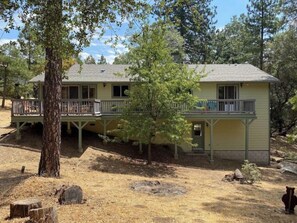 A two-story house with a porch and a deck, surrounded by trees and dry grass.