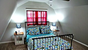 A bedroom with a sloped ceiling, a bed with a black metal frame, two bedside tables with lamps, and a window with red curtains. - The Wright Place (Kingsport)