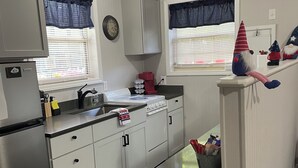 A compact kitchen with white cabinets, a stainless steel refrigerator, a stove, and a sink. There are two windows with blue curtains, a clock on the wall, and a small trash bin. - The Wright Place (Kingsport)
