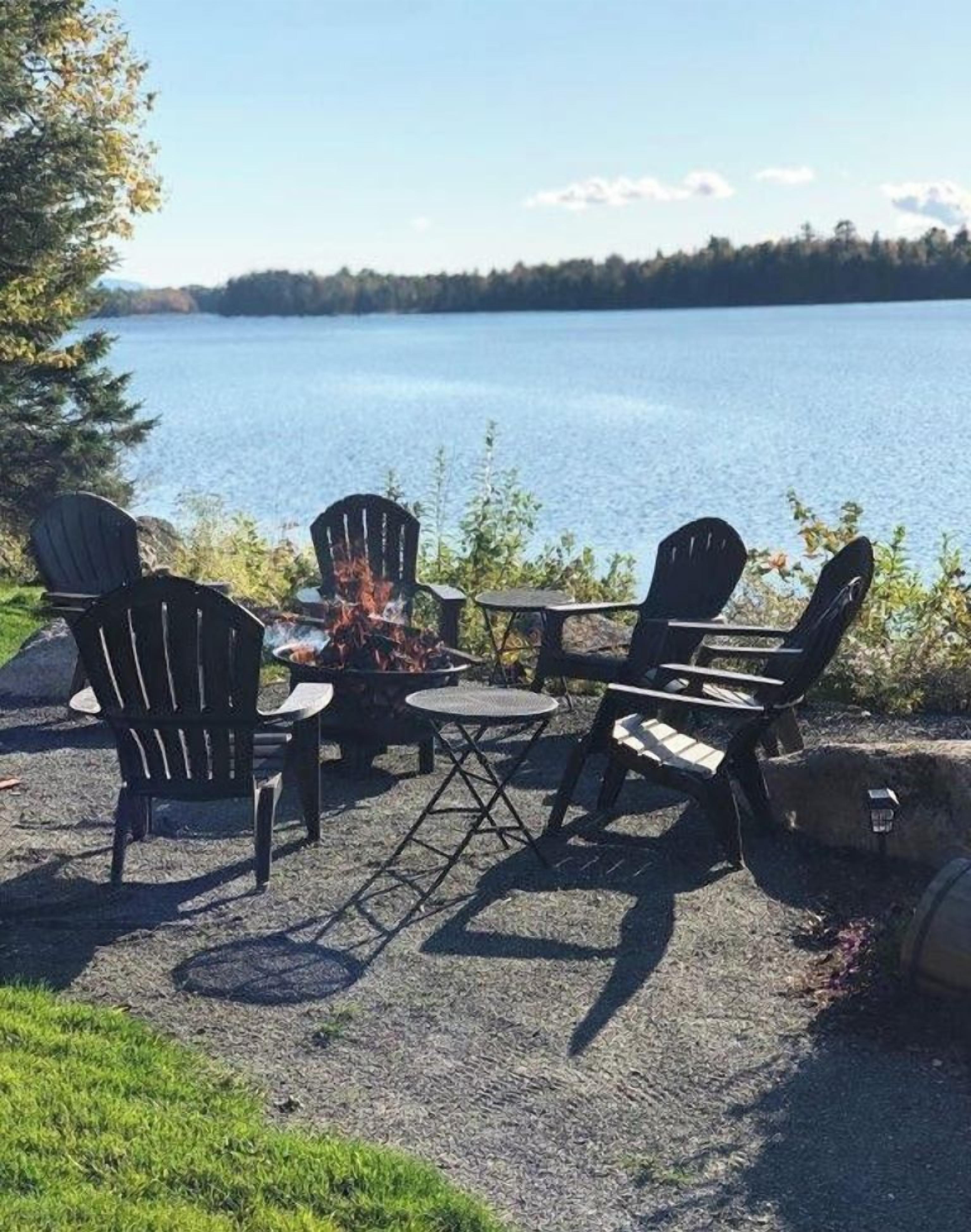 An outdoor seating area with Adirondack chairs and a small table, overlooking a lake.