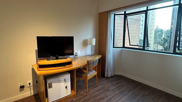 A hotel room with a flat-screen TV, wooden desk, chair, and a large window with a city view. - Cozy apartment with modern design - Sol Alphaville Hotel & Residence (Barueri)