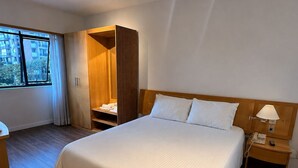 A hotel room with a large bed, a wooden wardrobe, a bedside table with a lamp, and a window with curtains. - Cozy apartment with modern design - Sol Alphaville Hotel & Residence (Barueri)