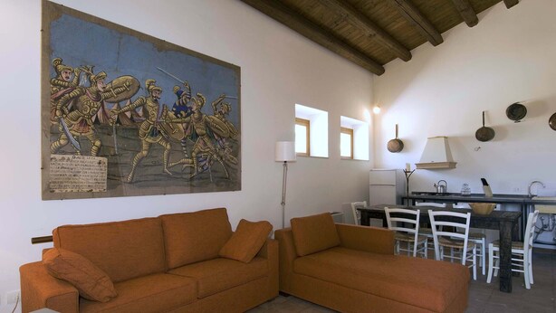 A living room with a large mural, a sofa, a dining table with chairs, and a kitchen area. - Nice apartment with private terrace. Shared pool. In Donnafugata, Sicily (Donnafugata)