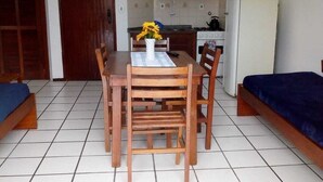 A compact room with a wooden dining table, chairs, a small kitchen area, and a bed with a blue cover.