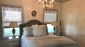 A bedroom with a bed, two bedside tables, a chandelier, and two windows with blinds. - SunShiner Farm - Shiner, TX (Shiner)