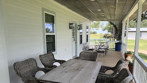 A covered porch with a wooden table and wicker chairs, a railing, and a view of a lawn and trees. - SunShiner Farm - Shiner, TX (Shiner)