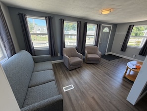 Ein Zimmer mit grauem Sofa, zwei Sesseln, einem kleinen runden Tisch und einem Fenster mit Vorhängen.