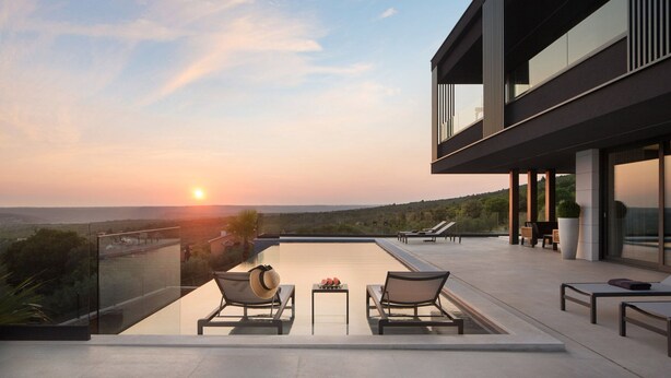 Zona de piscina exterior moderna con sillones de descanso y balaustrada de vidrio con vistas a un paisaje pintoresco al atardecer.