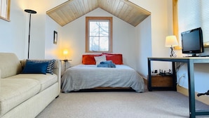 A bedroom with a sloped ceiling, a bed with red and blue pillows, a desk with a computer, and a TV.