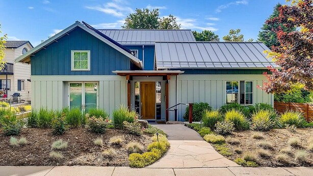 A two-story house with a blue exterior and a grey roof, featuring a front porch and a landscaped garden.