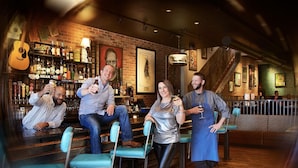 A bar with wooden walls, a brick fireplace, and a variety of liquor bottles. There are bar stools and a staircase leading to an upper level.