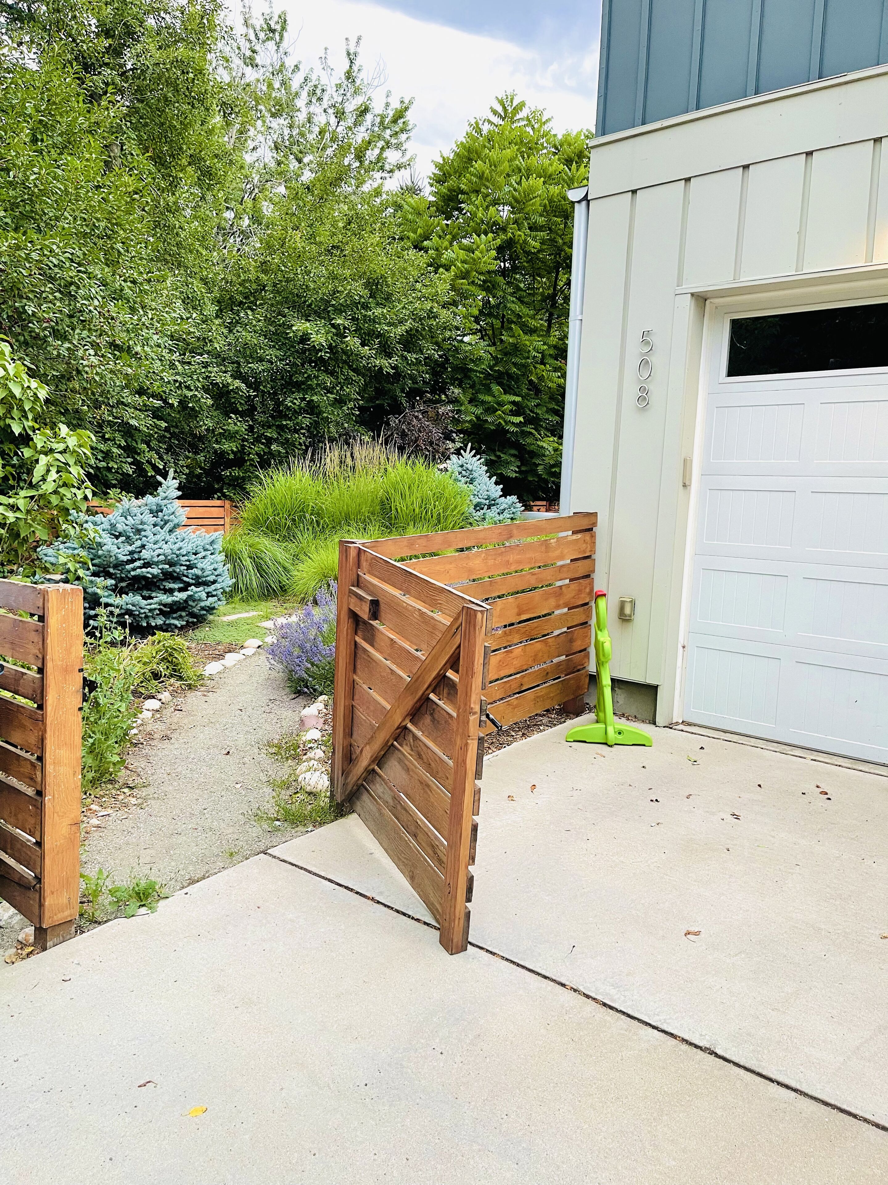 A wooden gate leading to a driveway, surrounded by lush greenery and a garage.