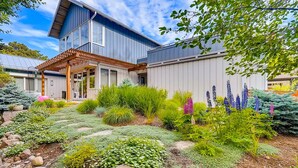 A modern two-story house with a covered patio, a landscaped garden with various plants and flowers, and a clear blue sky.