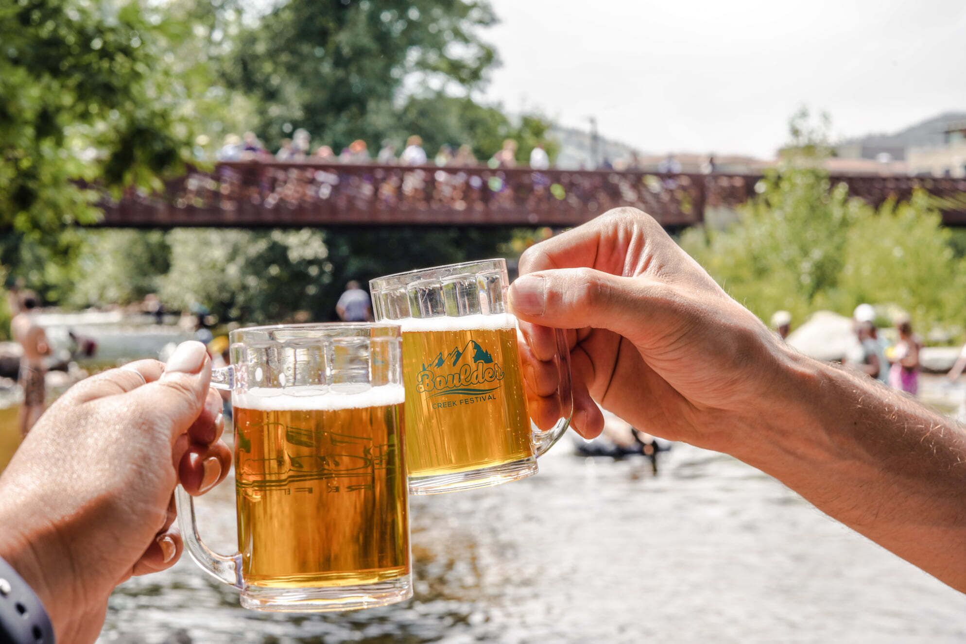 Zwei Hände halten Bierkrüge, im Hintergrund eine Brücke und Menschen.