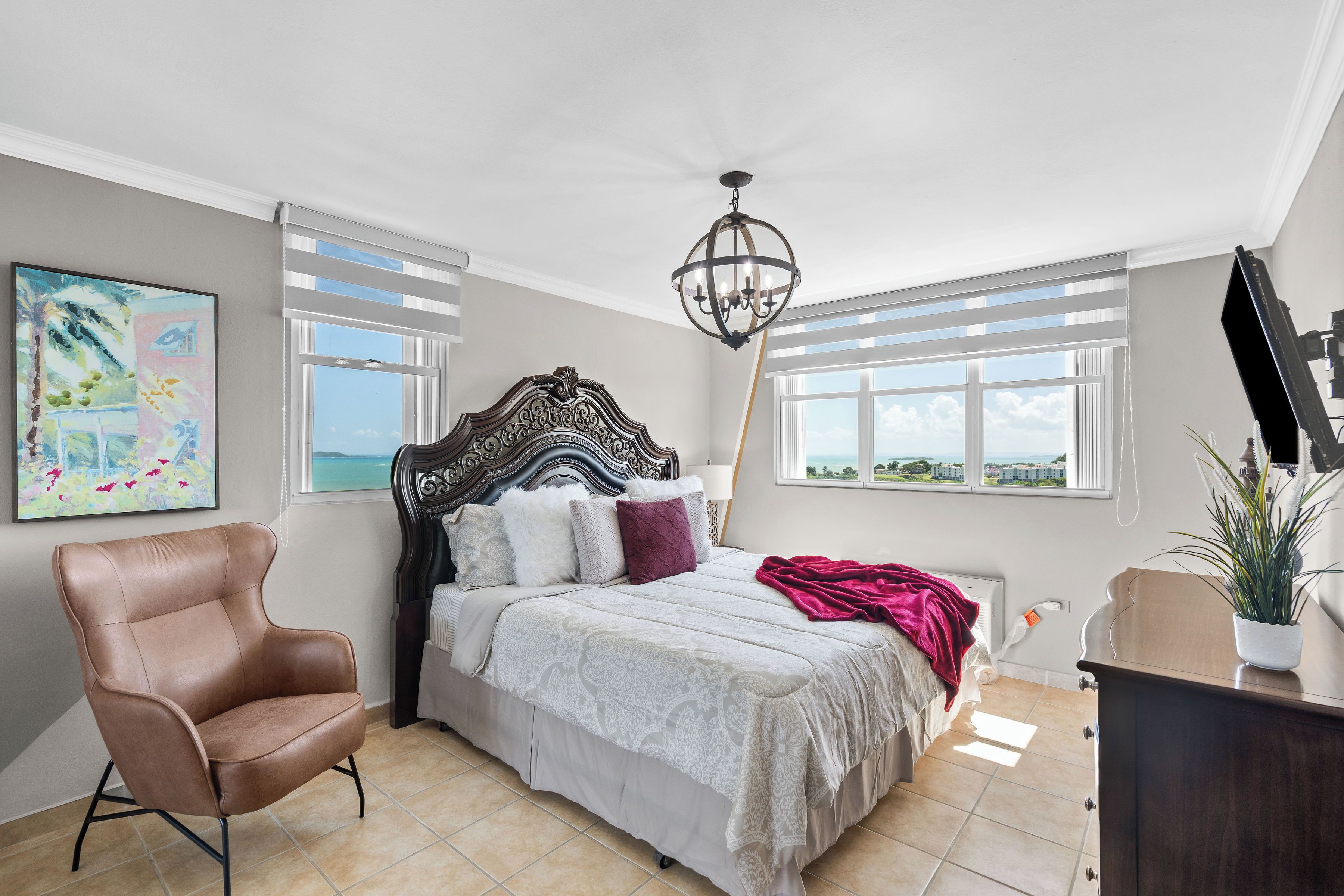 A bedroom with a bed, a chair, a chandelier, and a view of the outdoors.