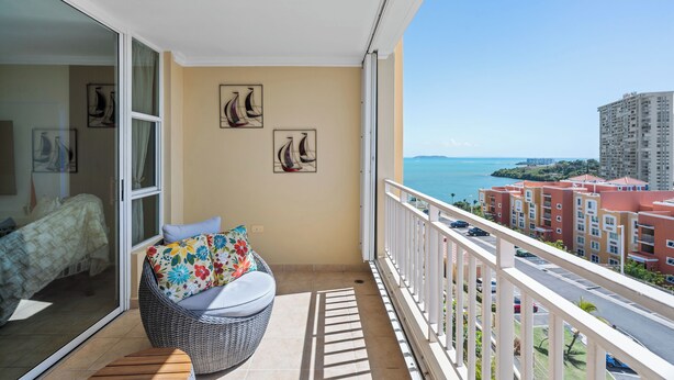 Ein Balkon mit einem Korbstuhl und Kissen, ein Bett mit Blumenmuster-Bettdecke und Blick auf das Meer und Gebäude.
