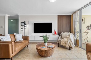 A modern living room with a leather sofa, a round wooden coffee table, a television mounted on the wall, and a large window with curtains.