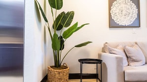 Un salon avec un canapé beige, une petite table d’appoint noire, une grande plante en pot et une œuvre d’art murale.