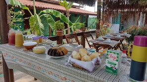 A table set for a meal with various food items and drinks, surrounded by wooden chairs and a green backdrop with plants. - Hawaii Flats (São Miguel do Gostoso)