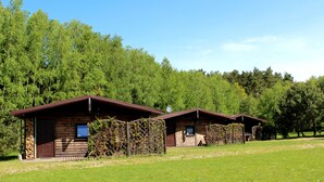 Dos cabañas de madera con techos marrones y paredes de madera, rodeadas por un campo cubierto de hierba y árboles frondosos.