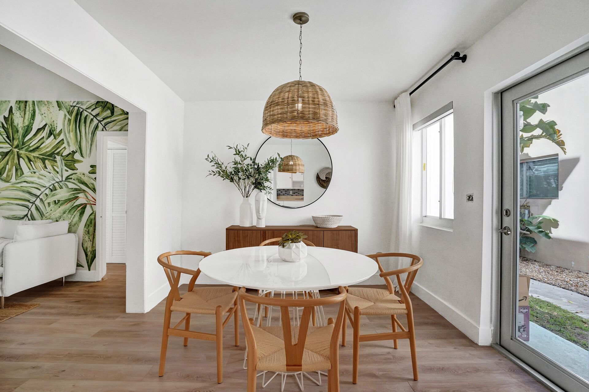 A dining area with a round table, four wooden chairs, a hanging woven light fixture, and a white vase with greenery.