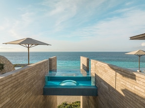 Piscina infinita moderna con vista despejada al océano, terraza de madera y dos sombrillas.