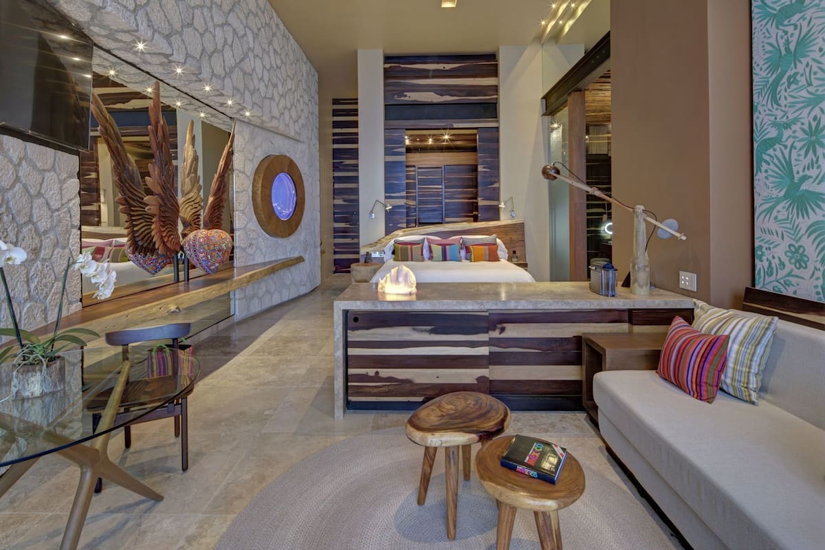 A modern hotel room with a stone wall, wooden accents, a central island, and a sofa with colorful pillows.