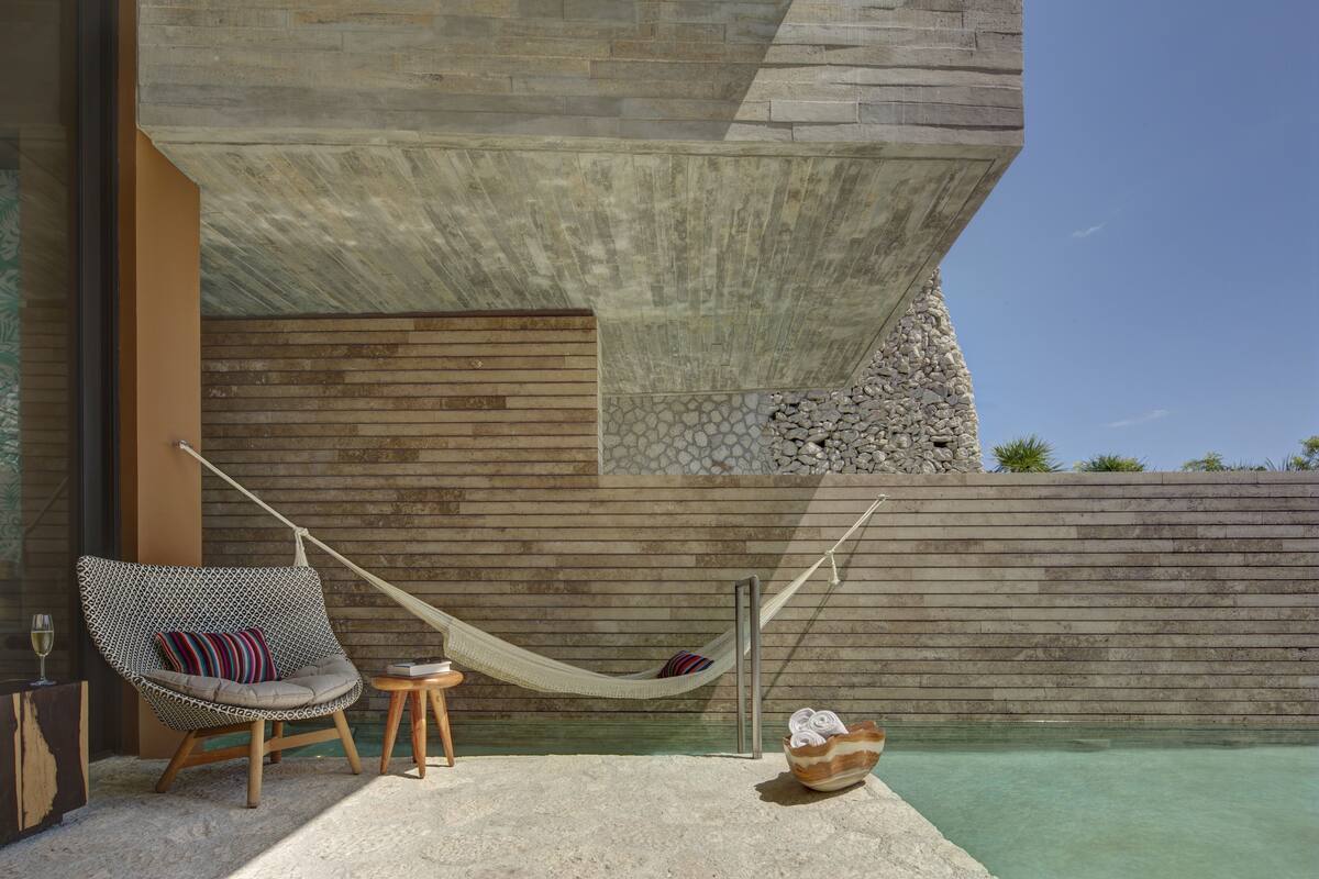 A hammock chair with a cushion, a small wooden stool, and a shell-shaped bowl by a pool with a stone wall backdrop.