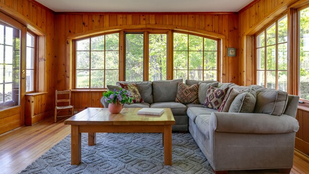 A cozy living room with wooden walls, a sectional sofa, a coffee table with a book titled "HORSE", and large windows offering a view of trees.