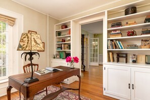A dining area with a wooden table, a lamp, and a vase with flowers. There are bookshelves with books and decorative items.