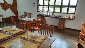 Un comedor con mesas y sillas de madera, pared de ladrillo con un hueco, y ventana con vista a la vegetación.