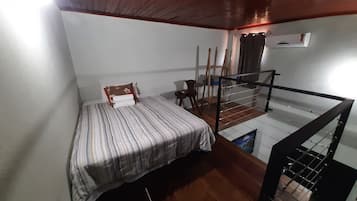 Una habitación con cama, techo de madera, una mesita, una silla y barandilla de vidrio.
