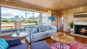 A modern living room with a sofa, armchairs, a glass coffee table, a fireplace, and a large window offering a view of the outdoors.