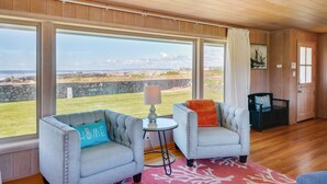 A spacious room with large windows offering a view of the beach and ocean. There are two upholstered chairs with cushions, a small round table with a lamp, and a wooden floor.