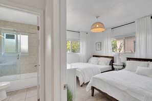 A bedroom with two beds, a bedside table, a window, and a bathroom with a shower and toilet.