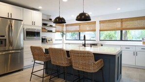 Een moderne keuken met een roestvrijstalen koelkast, een inbouwoven, een groot kookeiland met barkrukken, en een raam met zonwering.