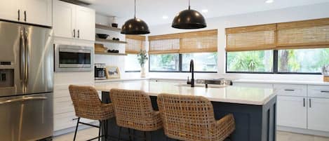 A modern kitchen with a stainless steel refrigerator, a built-in oven, a large island with bar stools, and a window with blinds.