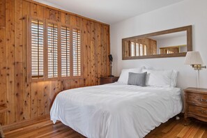 A bedroom with a wooden paneled wall, a large mirror, a bed with white bedding, a nightstand, and a lamp. - Moonridge Mountain Retreat! (Big Bear Lake)