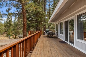 A wooden deck with outdoor furniture, surrounded by tall trees, and a house with large windows. - Moonridge Mountain Retreat! (Big Bear Lake)
