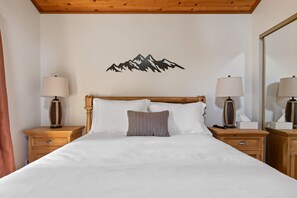 Ein ordentlich bezogenes Bett mit weißen Leinen, zwei Nachttische mit Lampen und ein an der Wand befestigtes Bergmotiv.