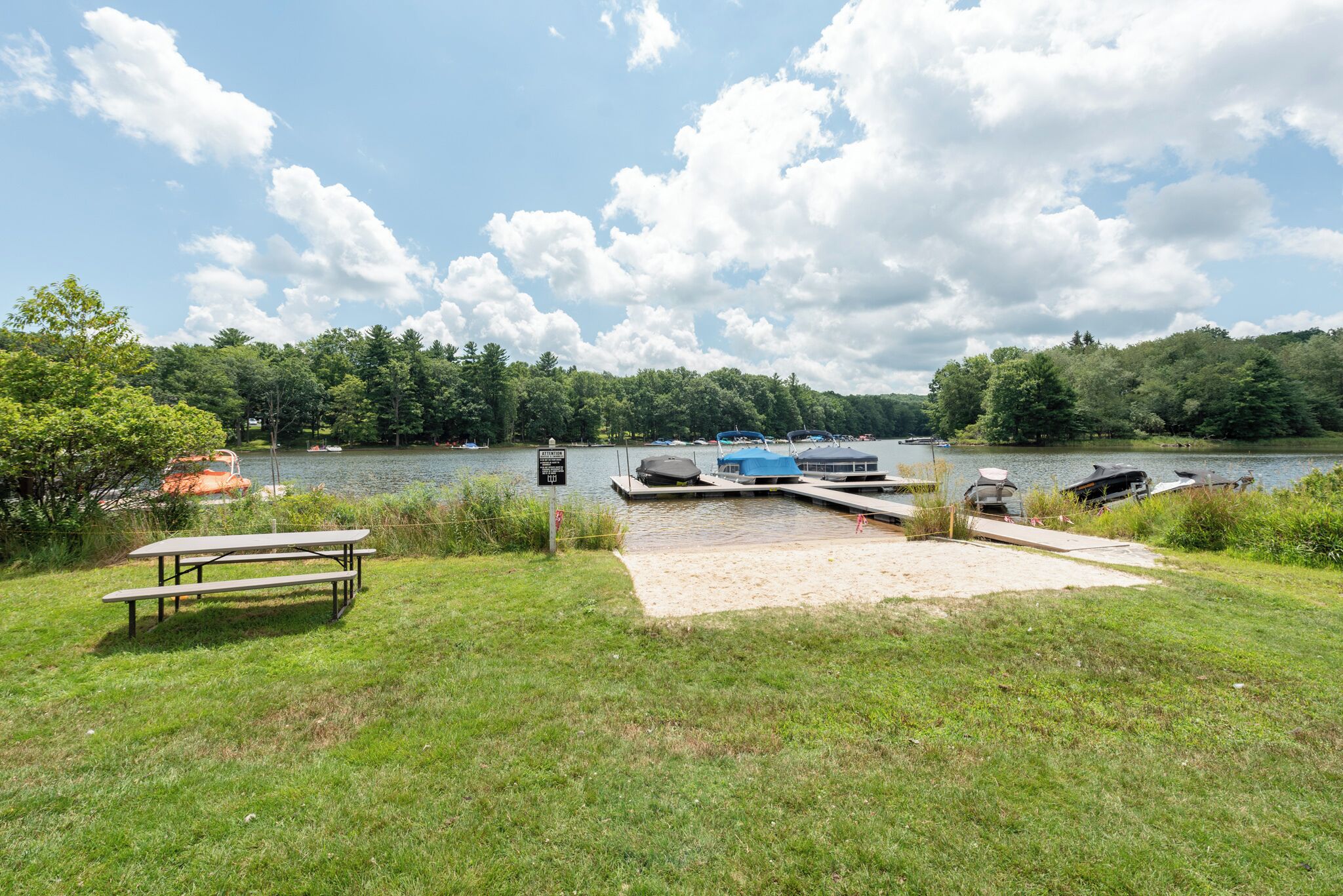 A lakeside picnic area with a bench, a dock with boats, and a sign indicating a boat launch area.