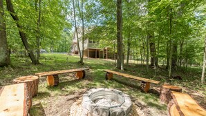 A rustic outdoor seating area with a stone fire pit, surrounded by wooden benches and a large house in the background. - Cozy Chalet w/ Lake Access & Game Room (Oakland)
