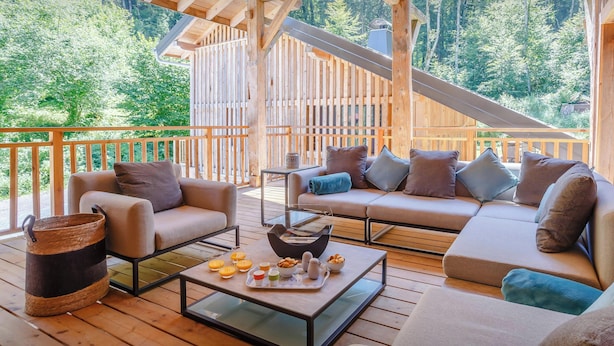 Une terrasse en bois avec un canapé et des fauteuils, une table basse avec des collations, et une vue sur une zone boisée.