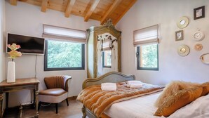 Une chambre avec un plafond en bois, un grand lit avec une tête de lit, une chaise, un bureau avec un vase et un téléviseur fixé au mur.