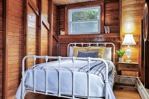 A wooden cabin bedroom with a metal bed frame, a bedside table with a lamp, and a window with a view of trees.